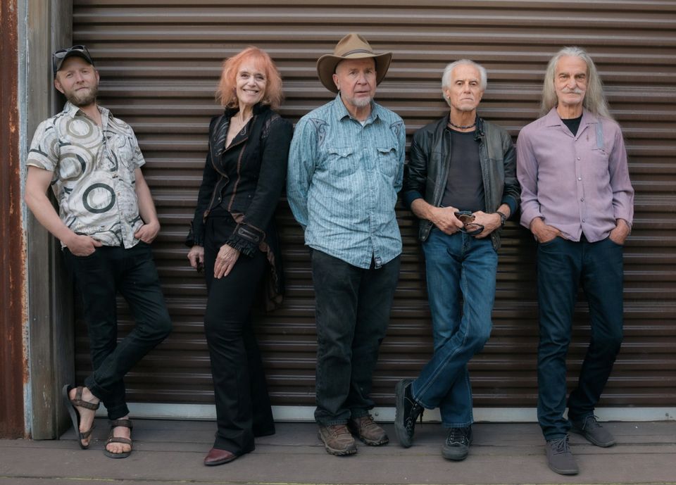 Photo of Deep Pockets band. From left to right A man leaning against a door frame, a woman leaning her back on a man wearing a hat, to the right is man holding his belt buckle, and on the far right a man is standing with pockets in his hands. They are in a band