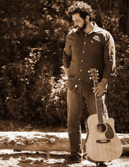 A sepia toned photo of Arron Ford holding a guitar looking at the ground.