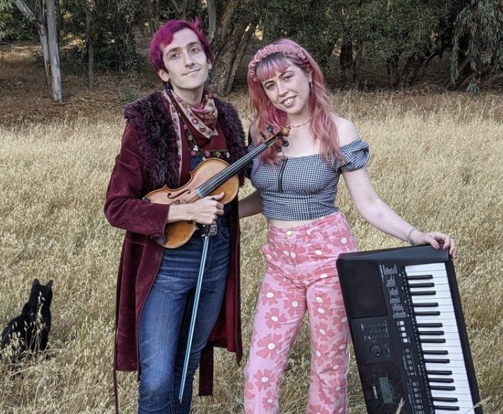 A picture of Corwin and Grace in a field on the edge of a oak forest. Corwin is holding a violin and Grace is holding a keyboard. They are embracing each other in an open hug.