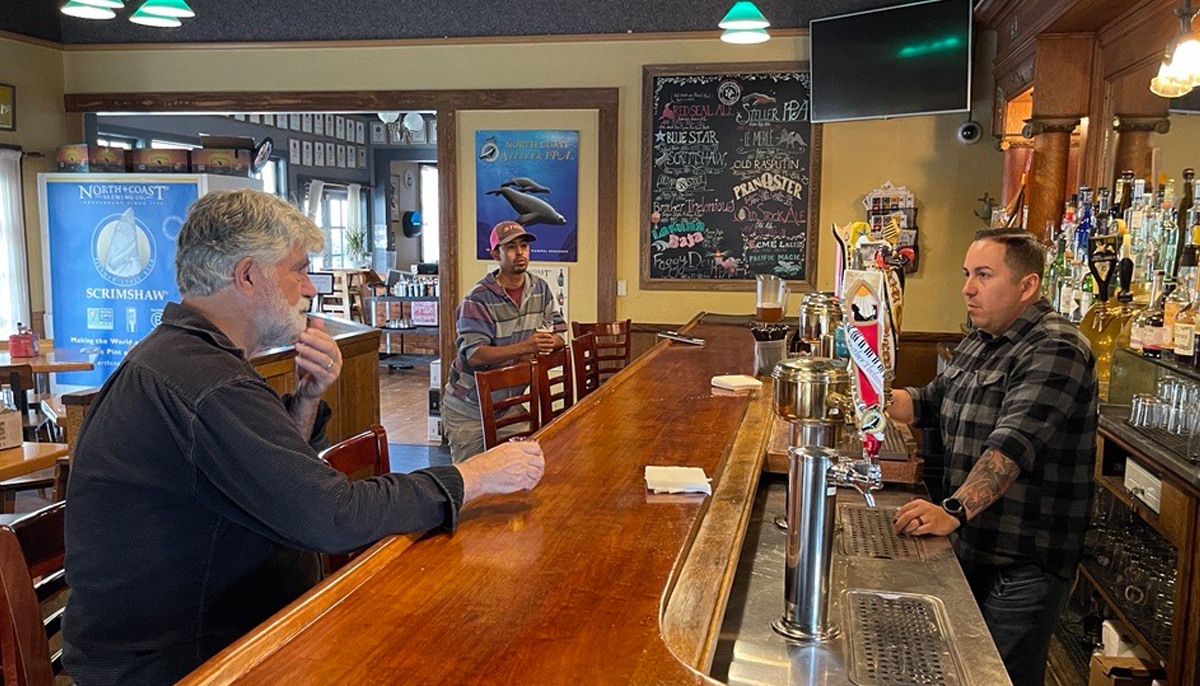 The assistant brewmaster, Obed Salazar, serves the original founding brewer, Mark Ruedrich, a sample of this years exclusive Festbier. Another brewer is seen in the background near a chalkboard menu and posters showcasing North Coast Brewing’s products .