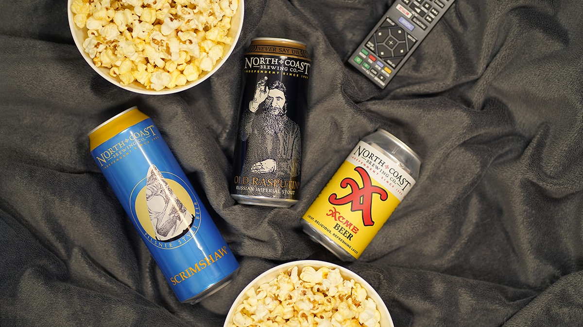 A 19.2oz can of Scrimshaw, 16oz can of Old Rasputin, and 12oz can of Acme Beer with two bowls of popcorn and a television remote on a grey blanket.