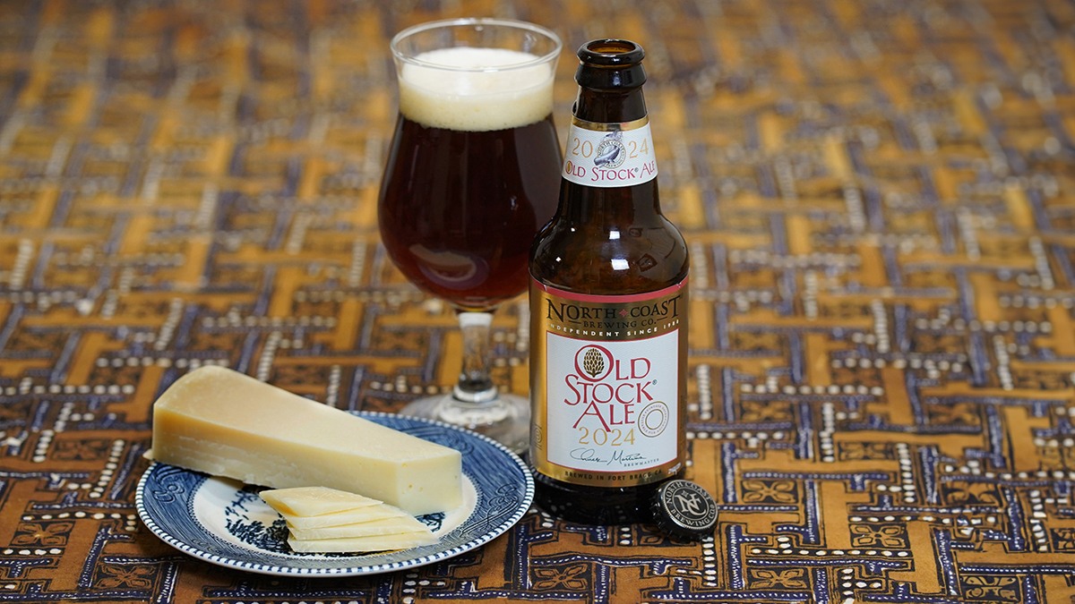 A 12oz bottle of Old Stock Ale next to a glass of beer and a plate with slices of Parmigiano Reggiano cheese on a brown tablecloth.