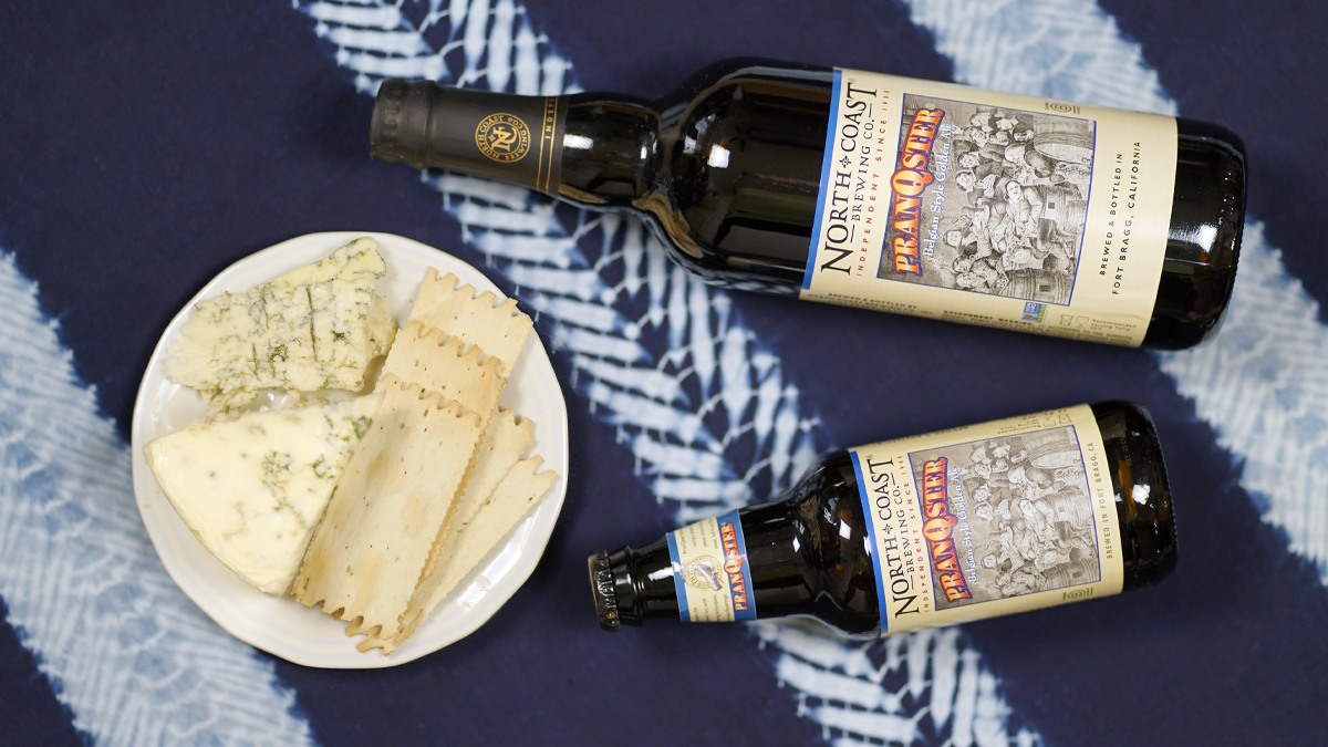 12oz and 750mL bottles of PranQster Belgian Style Golden Ale laying next to a plate with blue cheese and crackers on a blue tablecloth.