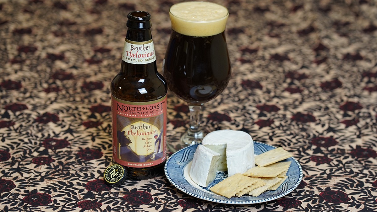 A 12oz bottle of Brother Thelonious Belgian Style Abbey Ale next to a glass of beer and a plate with a small brie cheese on a dark floral tablecloth.