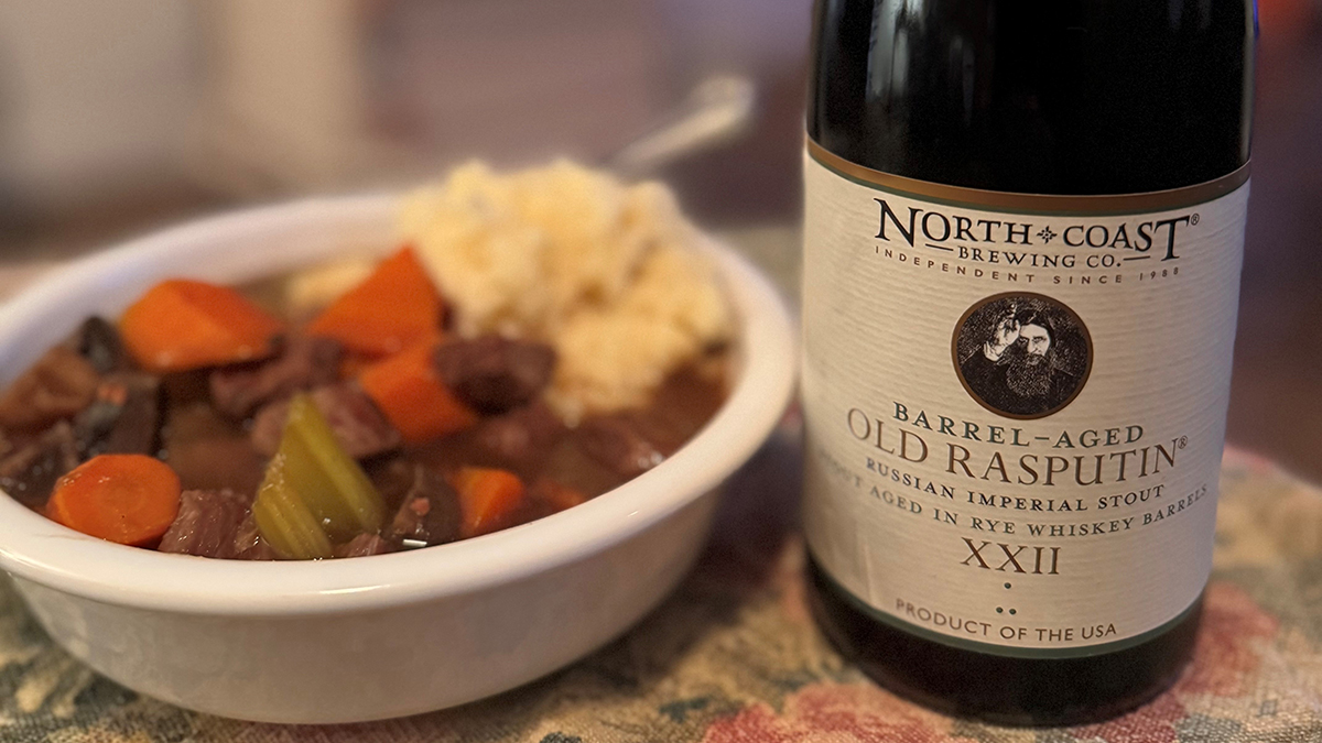 A bowl of beef stew next to a bottle of Barrel-Aged Old Rasputin Russian Imperial Stout on a table.