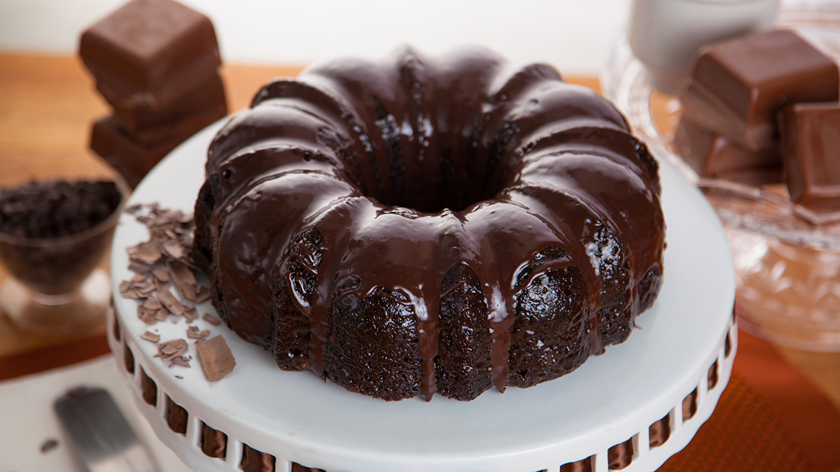 A dark chocolate bundt cake sitting on a white cake stand, surrounded by chocolate.