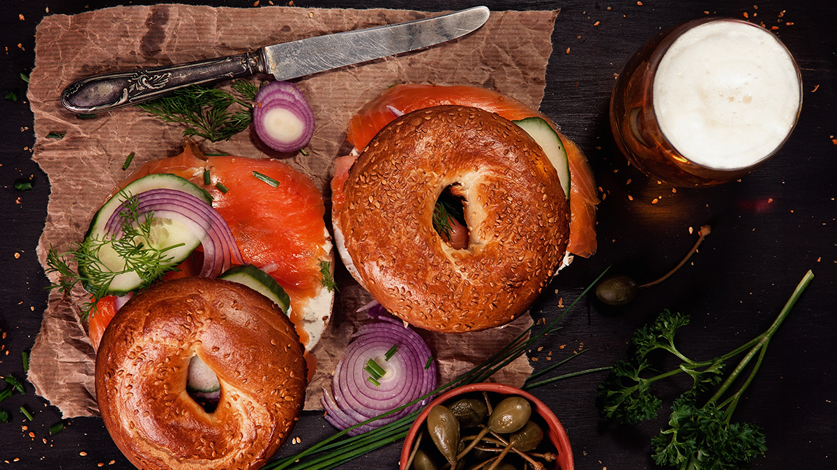 Top down view of two bagels with cream cheese, smoked salmon, cucumbers and red onion, next to a glass of beer.