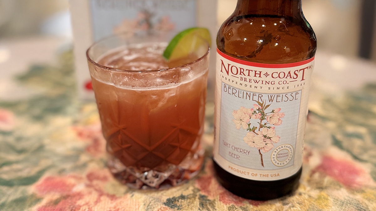: Tart Cherry Gin Rickey garnished with a lime wedge, next to a bottle of Tart Cherry Berliner Weisse, on a floral tablecloth.