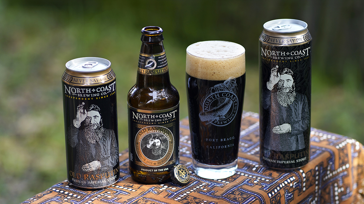 16oz can, 12oz bottle and 19.2oz can od Old Rasputin sit next to a glass of stout on a bench outdoors.