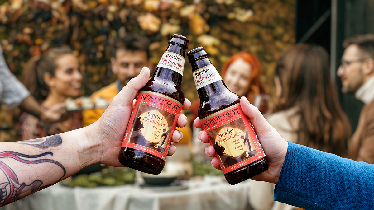 Two hands toasting with 12oz bottles of Brother Thelonious in front of a fall dinner celebration.