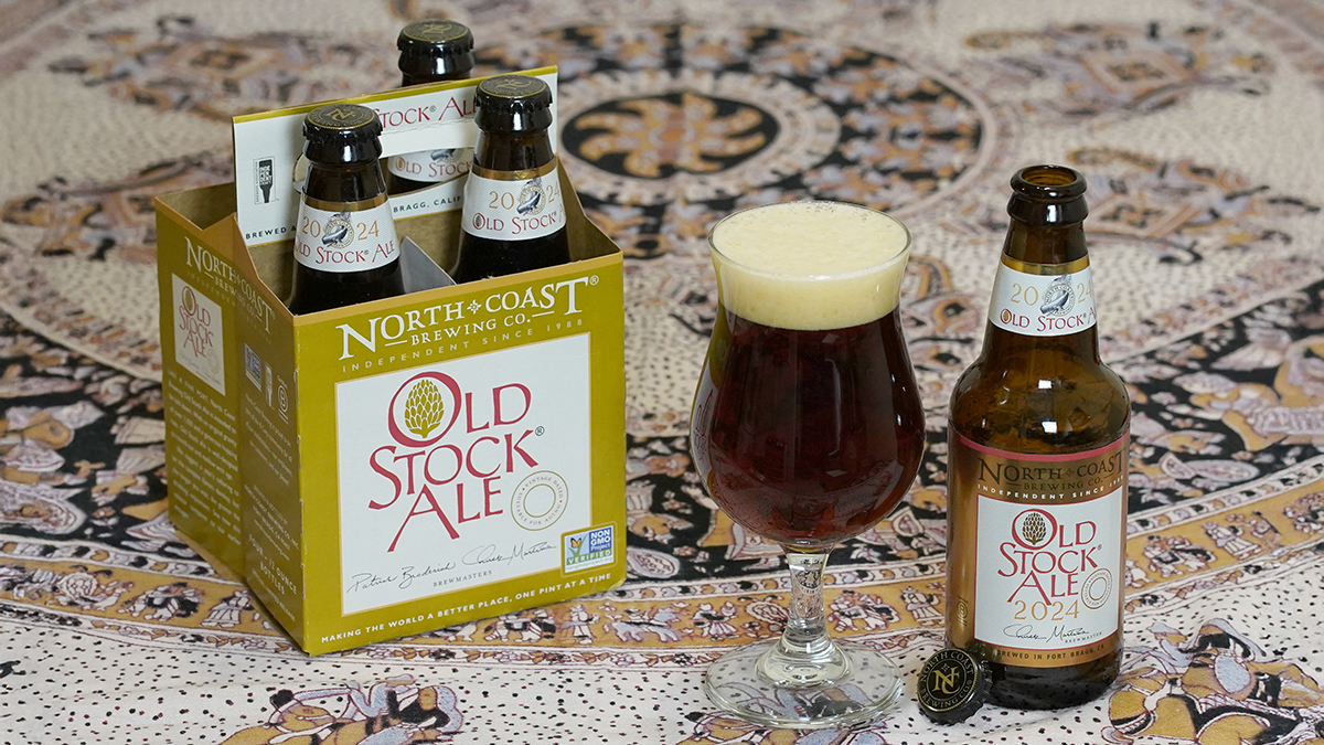 A glass of Old Stock Ale next to an empty bottle and a four pack of bottles sitting on a lively tablecloth.