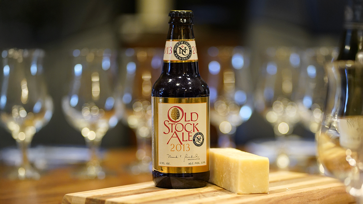A bottle of Old Stock Ale sitting next to a block of cheese on a cutting board with many glasses waiting in the background.