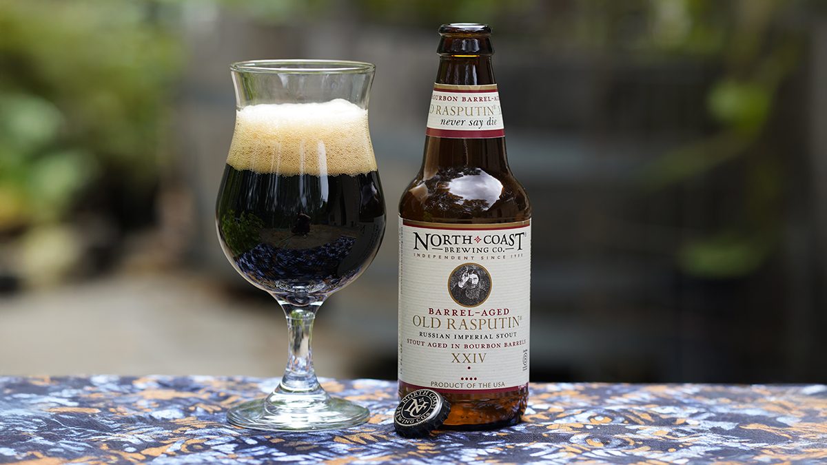 12oz bottle of Barrel-Aged Old Rasputin XXV sits next to a glass of beer on a table outdoors.
