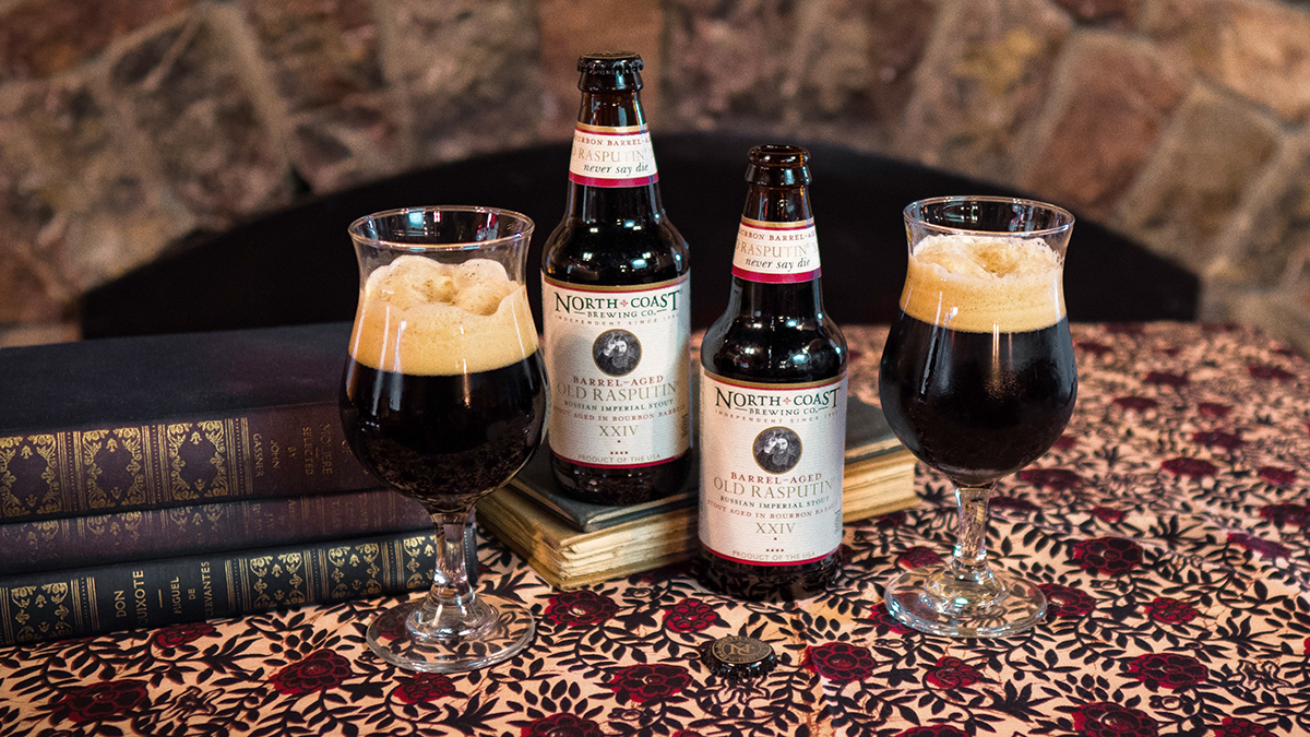 Two 12oz bottles of Barrel-Aged Old Rasputin XXIV with glasses of beer on a table in front of a fireplace