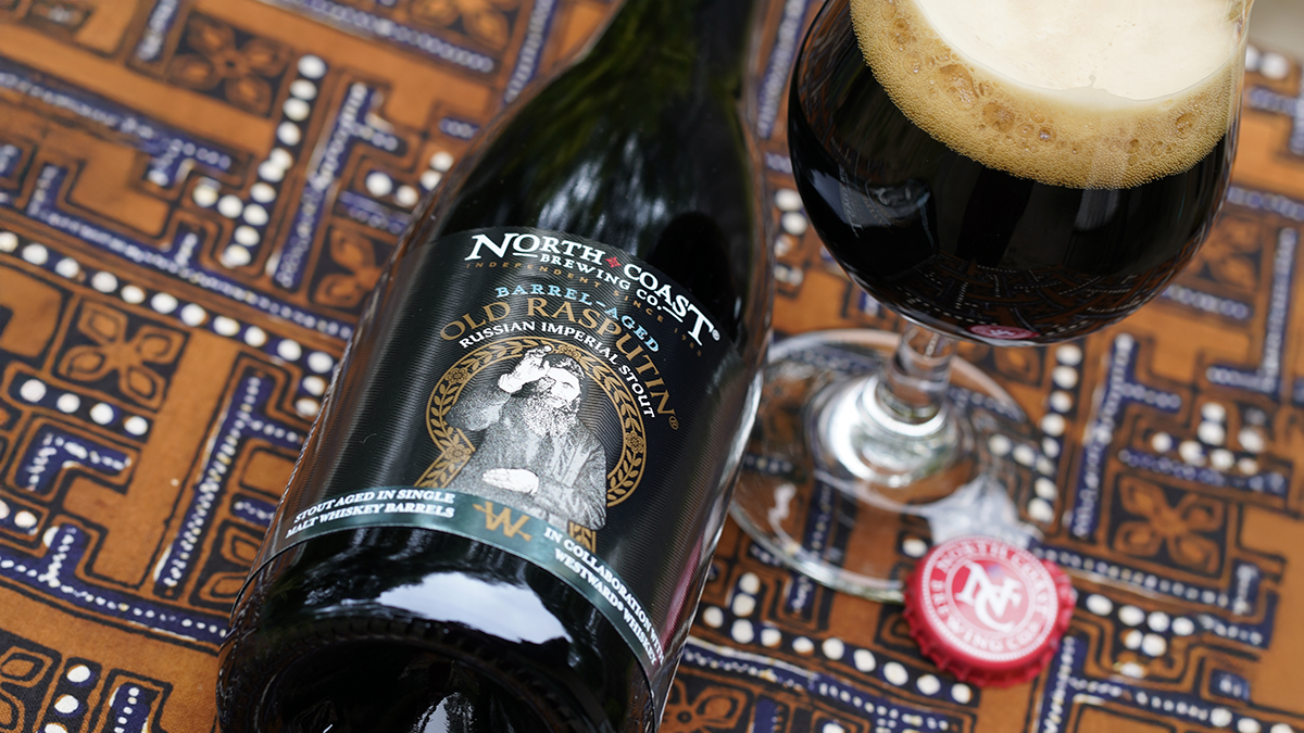 Top view of a 500ml bottle of Barrel-Aged Old Rasputin laying next to a glass of beer on a brown tablecloth.