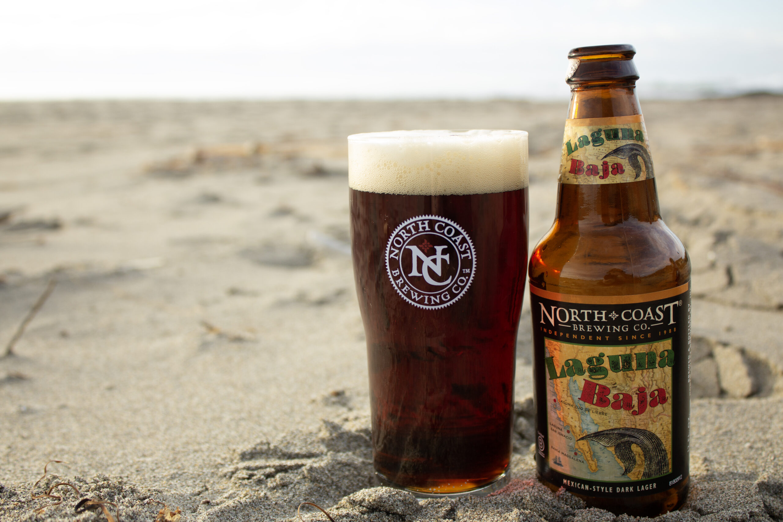 Empty bottle of Laguna Baja next to a full glass of beer on the beach.