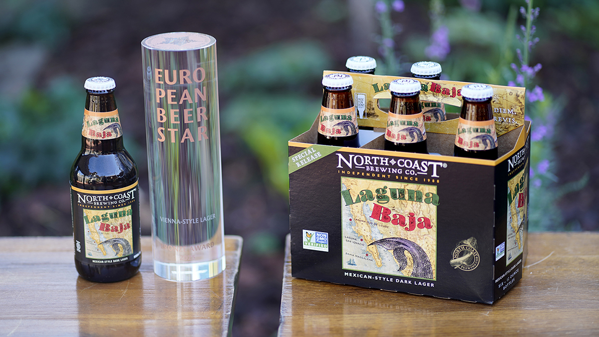 Laguna Baja six-pack and single bottle next to European Beer Star glass trophy on two tables outdoors.