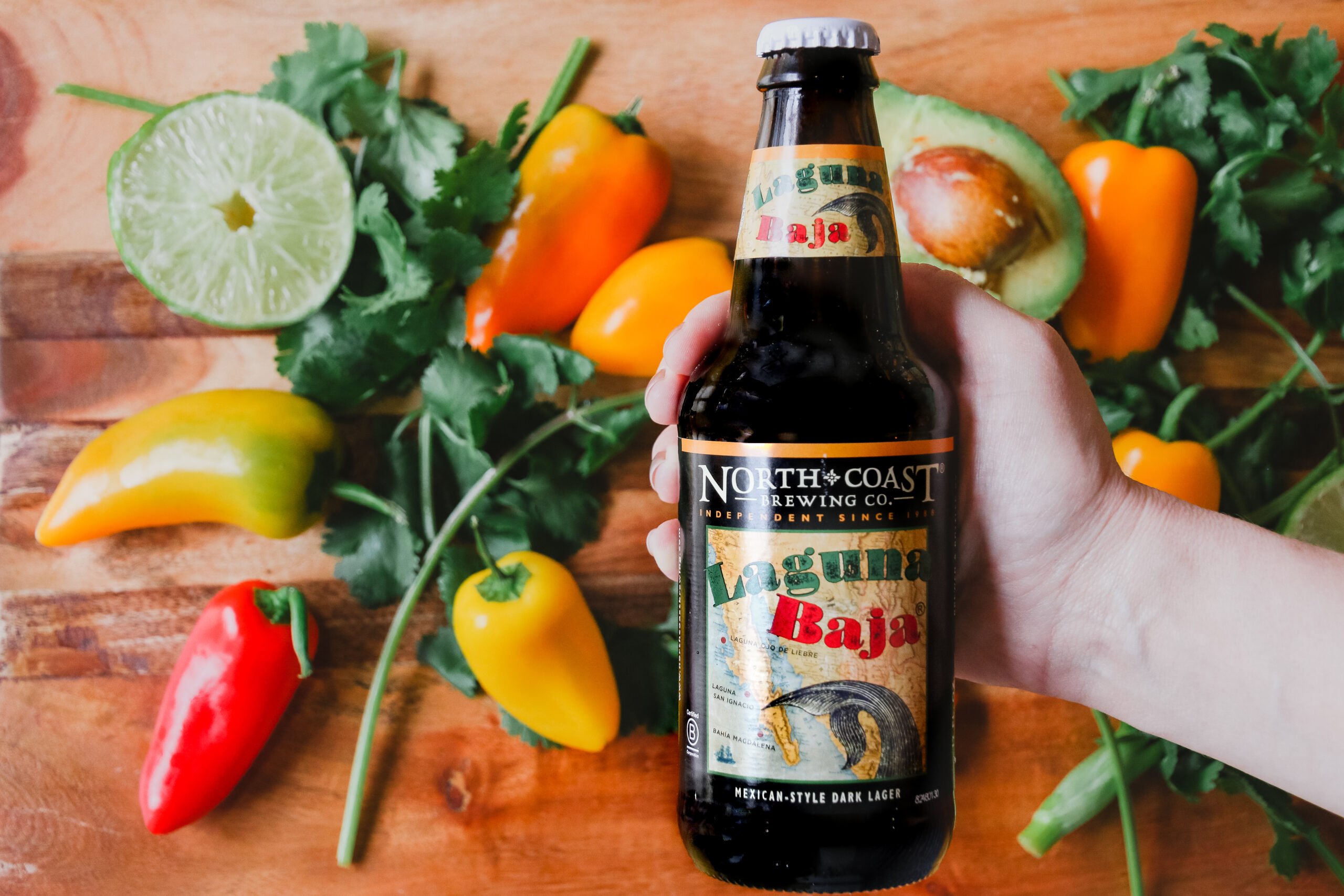 A hand holds a bottle of Laguna Baja above a cutting board with peppers, avocado, cilantro, and lime.