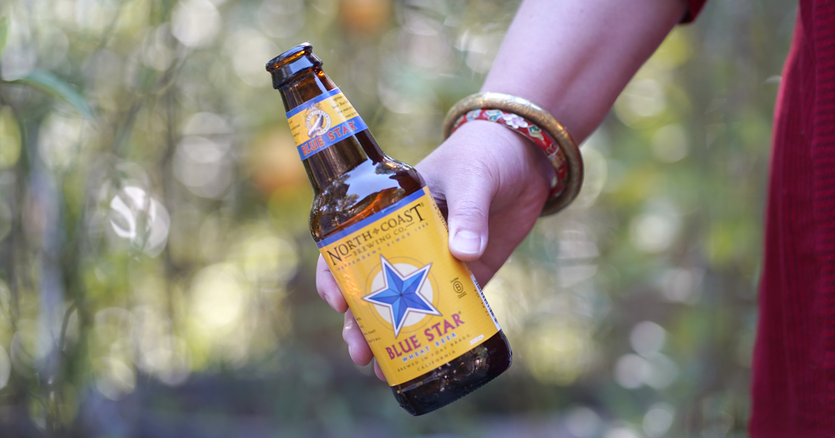 A hand holds a bottle of Blue Star wheat beer outdoors.
