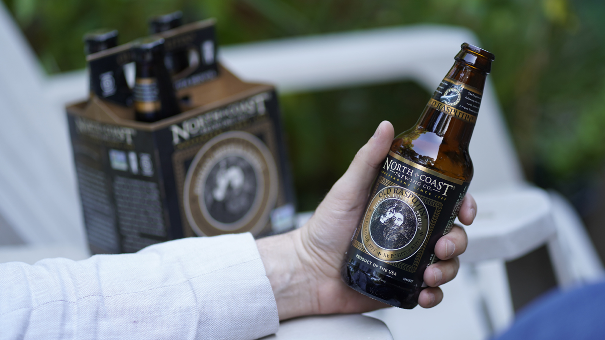A man's hand holds a bottle of Old Rasputin while sitting in a backyard lawn chair, with the four pack carrier in the background.
