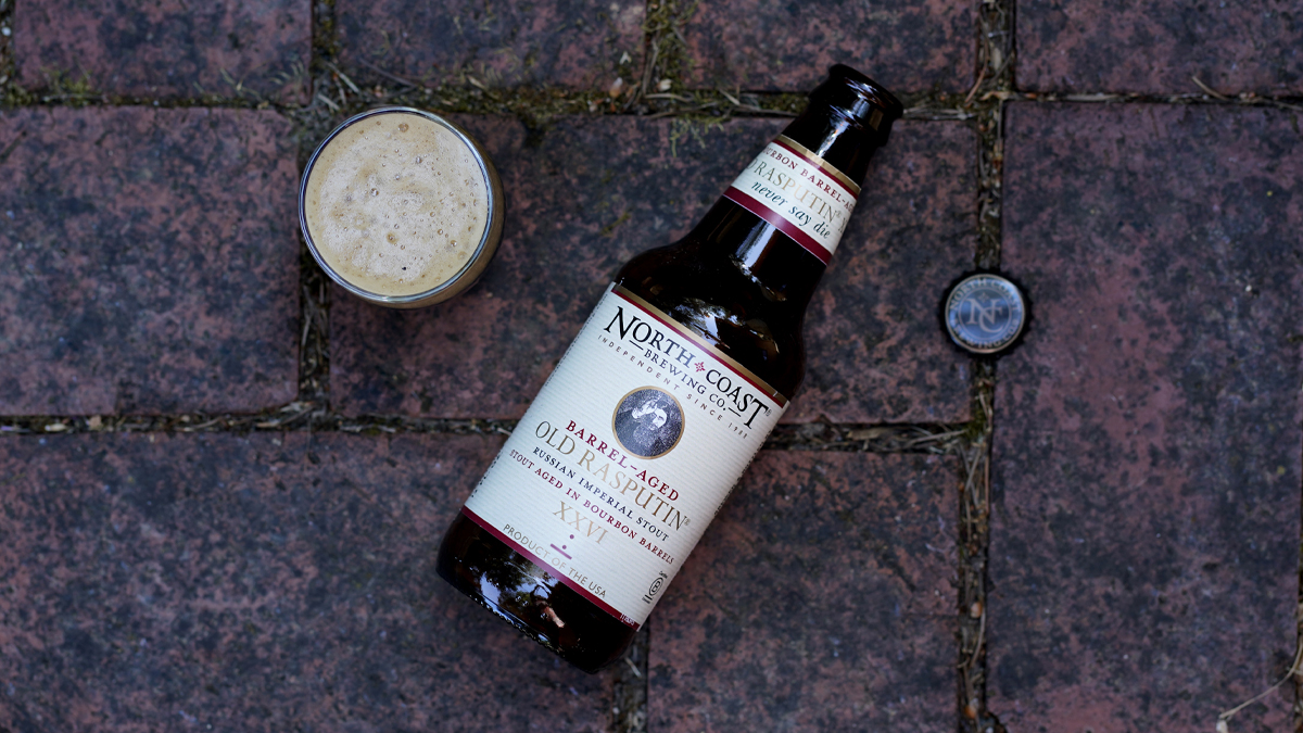 A bottle of Barrel-Aged Old Rasputin lays on a brick background, next to a taster glass of beer.
