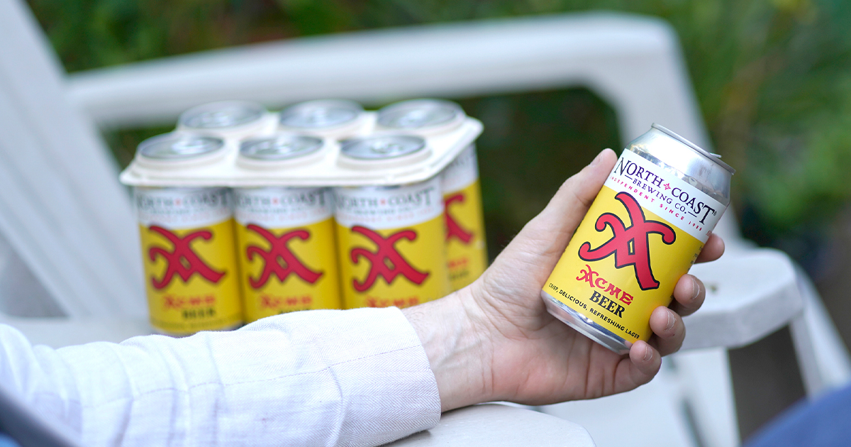 A man's hand holds a can of Acme Beer while sitting in a lawn chair, with a 6-pack of Acme Beer on the chair beside him.