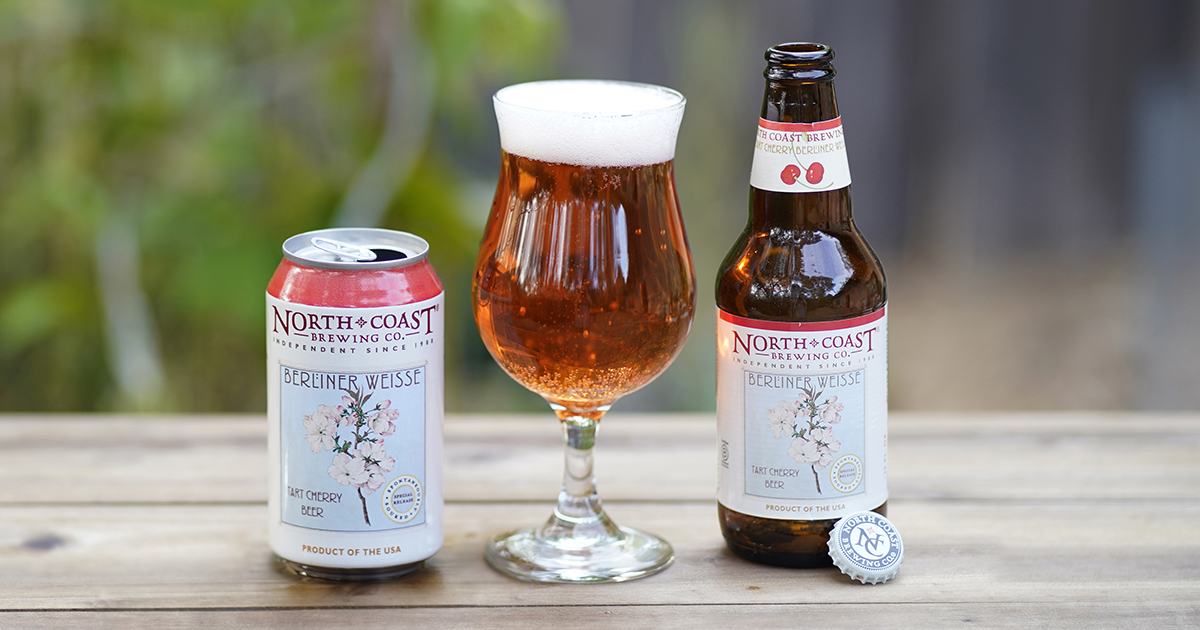 Open can and bottle of Tart Cherry Berliner Weisse next to a glass of the beer on a picnic table outdoors.