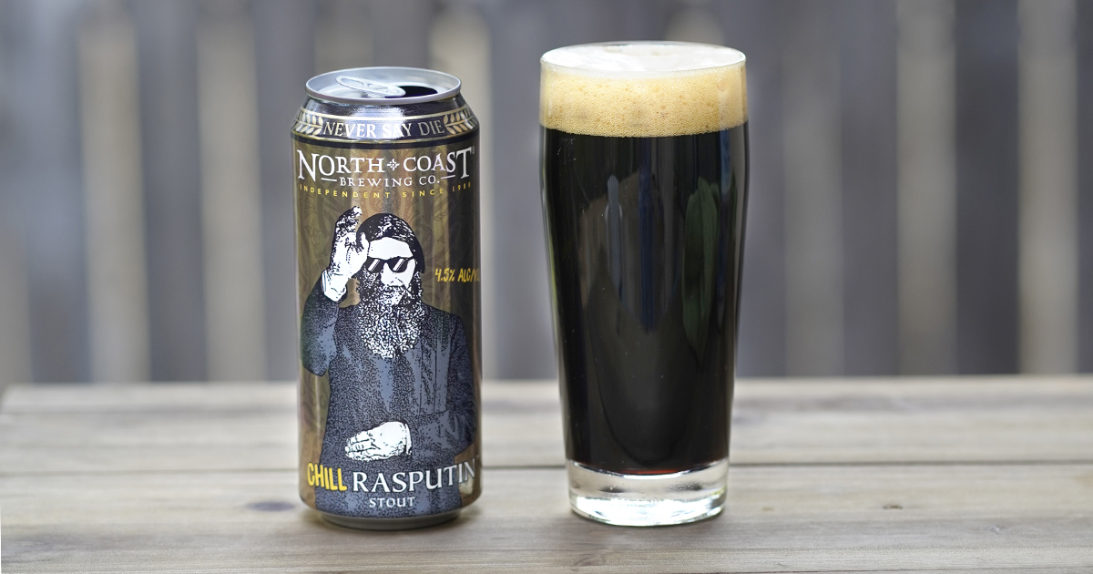 Open can of Chill Rasputin next to a glass of the stout on a picnic table outdoors.