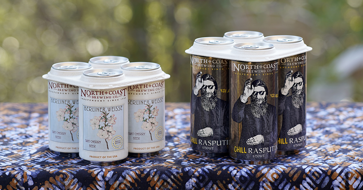 A four pack each of Tart Cherry Berliner Weisse cans and Chill Rasputin cans sit on a colorful tablecloth outdoors.